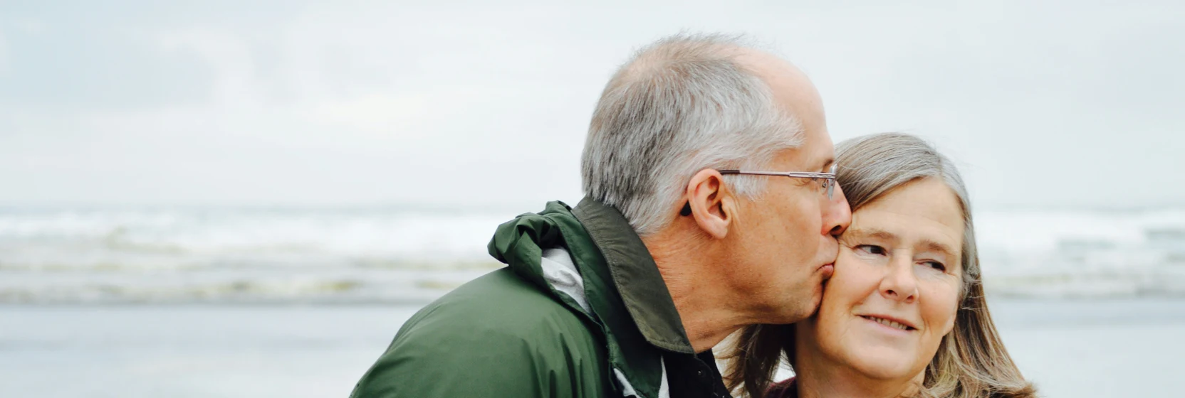 A couple in their sixties on a beach, one kissing the other on the cheek